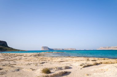 Geniş soluk kum düzlükleri Girit 'in Balos Sahili' nde berrak, bulutsuz bir gökyüzü altında kayalık adalar ve uzak kayalıklara uzanan canlı mavi bir lagüne doğru uzanır. Manzara güneşli, ferah ve huzurlu..