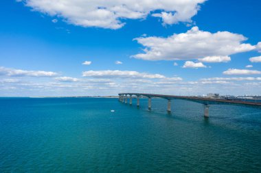 Karayı Charente-Maritime 'deki Ile de Re' ye bağlayan, dağınık kümülüs bulutlarıyla dolu bir gökyüzünün altında, canlı mavi su boyunca uzanan uzun beton köprü. Açık deniz manzarası sakin kıyı ortamını ve ferah atmosferi vurgular.