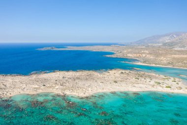 Geniş bir kumlu kıyı şeridi, canlı turkuaz sığ ve berrak mavi gökyüzü altında engebeli araziye sahip Elafonisi, Girit 'in hava manzarası. Manzara açık ve güneşli, sakin bir Akdeniz atmosferini çağrıştırıyor..
