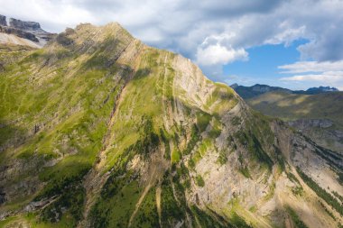 Değişen bulutların altında, Pireneler 'de dramatik kaya oluşumları ve güneş ışığı parçalarıyla sarp, çimenli dağ yüzeyi. Engebeli dokular ve keskin hatlar vahşi alp manzarasını vurgular.