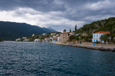 Renkli çatıları olan geleneksel evler, sakin bir sahil köyünün kayalık kıyı şeridi, ön planda derin mavi sular ve karamsar, bulutlarla dolu bir gökyüzünün altında yükselen engebeli dağlar..