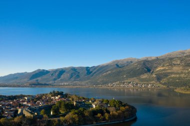 Arka planda engebeli Pindus dağlarının yükseldiği Ioannina Gölü ile çevrili tarihi Ioannina yarımadasının havadan görünüşü. Sahnede ortaçağ duvarları, kırmızı kiremitli çatılar ve Epirus 'taki canlı mavi gökyüzünün altında sakin bir su yer alıyor.