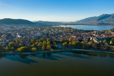 Gökyüzü perspektifi, Ioannina Gölü ile çevrili Ioannina şehrinin kırmızı kiremitli çatıları, yemyeşil ağaçları ve arka planda yükselen Pindus dağları olduğunu ortaya koymaktadır. Sahne, berrak bir gün ışığı, açık mavi gökyüzü ve huzurlu bir şehir manzarası içeriyor.