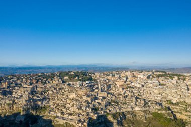 Matera, İtalya 'nın geniş hava görüntüsü engebeli arazide uzanan yoğun bir tarihi taş bina kümesini ufuktaki uzak dağları ve yukarıdaki parlak mavi gökyüzünü gösteriyor..