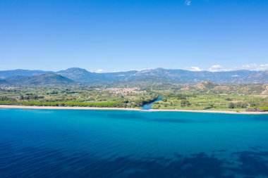Sardunya 'daki açık mavi gökyüzünün altında Lotzorai' yi çevreleyen yemyeşil ve uzak dağlarla dolu uzun kumlu bir plajla canlı bir turkuaz deniz manzarası..