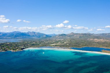 Yüksek açılı Spiaggia Lu Impostu manzarası canlı turkuvaz suları, beyaz kumlu kıyı şeridi ve bulutlarla benekli açık mavi gökyüzünün altındaki dağ arkaplanı..