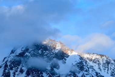 Sıcak güneş ışığı Aiguille du Midi 'nin engebeli, karlı zirvesini aydınlatır. Yumuşak bulutlar mavi gökyüzünde sürüklenirken sakin bir sabah atmosferi yaratır..