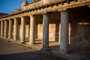 Ilık güneş ışığı Pompeii, İtalya 'daki Stabian Hamamları' nda eski Roma sütunları ve yıpranmış freskli duvarları aydınlatıyor. Kalıplı taş ve terracotta çatısı uzun gölgeler ve açık mavi gökyüzü tarafından vurgulanır..