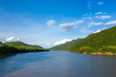 Parlak güneş ışığı geniş Mekong nehrini aydınlatır. Yoğun ormanlık tepeler arasında dalgalanan parlak mavi gökyüzü ve dağınık bulutlar. Manzarada verimli tropikal bitki örtüsü, yumuşak yamaçlar ve Güneydoğu Asya 'da sakin sular yer alıyor..