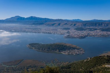 Ioannina 'nın sakin göl sularına kurulmuş ağaçlarla kaplı adasının geniş manzarası, Ioannina şehri ve Pindus dağları dramatik bir zemin oluşturuyor. Kıtır mavi gökyüzü ve yumuşak sabah sisi doğal ve kentsel manzarayı vurguluyor