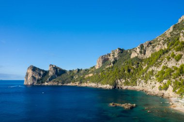 Dik uçurumlar ve yemyeşil bitki örtüsüyle kayalık kıyı şeridi bulutsuz bir gökyüzünün altında derin mavi Akdeniz sularıyla buluşuyor. Güneş ışığı, bu sakin İtalyan deniz manzarasının doğal dokularını ve canlı renklerini vurgular.