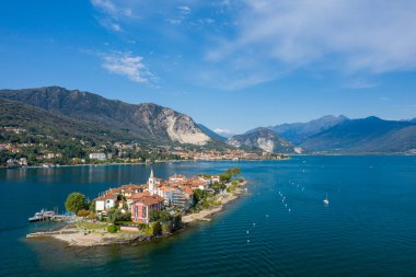 Isola dei Pescatori 'nin renkli binaları, kilise kulesi ve gür yeşilliğiyle Maggiore Gölü' nün derin mavi sularına karşı geniş bir hava perspektifi. Alp dağları ve göl köyleri açık bir gökyüzünün altında dramatik bir zemin oluştururlar.