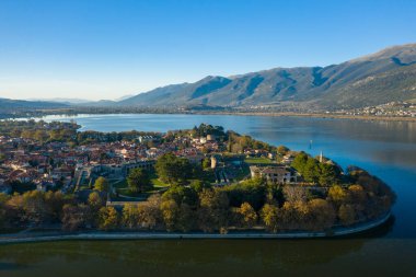 Geniş bir hava perspektifi, Ioannina Gölü ile çevrili tarihi Ioannina yarımadasını ve arka planda Pindus dağlarının yükseldiğini gösteriyor. Sahnede ortaçağ duvarları, kırmızı kiremitli çatılar ve berrak mavi gökyüzünün altında sakin bir su yer alıyor.