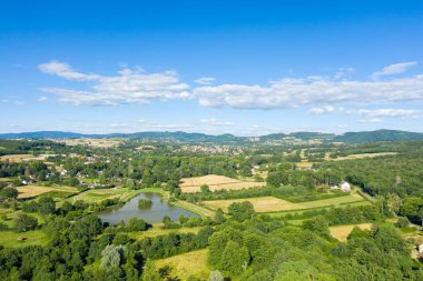 Yansıtıcı gölet, yamalı tarlalar ve Saint-Honore-les-Bains yakınlarında yemyeşil evlerle çevrili, parlak mavi bir gökyüzü altında, dağınık bulutlarla dolu, kırsal bir araziye geniş drone perspektifi..
