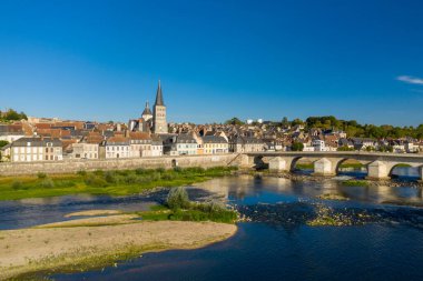 Parlak güneş ışığı tarihi taş köprüyü, kilise kulesini ve Loire nehri ve önde yemyeşil nehir yatağı bulunan La Charite sur Loire 'ın geleneksel evlerini aydınlatır. Açık mavi gökyüzü ve canlı renkler sakin atmosferi çağrıştırıyor