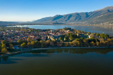 Ioannina Gölü ile çevrili tarihi Ioannina yarımadasının havadan görünüşü, kırmızı kiremitli çatılar, ortaçağ duvarları ve Pindus dağlarının arka planına kurulmuş verimli ağaçlar. Sahnede açık mavi gökyüzü ve sıcak öğleden sonra güneş ışığı var.