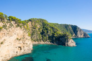 Bereketli yeşil çam ağaçları ile kaplı dik kayalık uçurumlar İyon kıyısı boyunca turkuaz deniz üzerinde yükseliyor. Parlak güneş ışığı ve bulutsuz bir gökyüzü taze, davetkar bir Akdeniz manzarası yaratır..