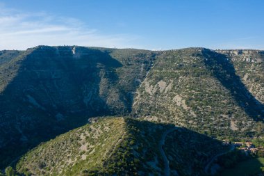 Geniş plato, açık mavi gökyüzü altında engebeli kireçtaşı yamaçları ve dağınık yeşil bitki örtüsüyle Navacelles Sirki 'ndeki derin bir kanyonun üzerinde yükselir. Güçlü güneş ışığı, dokulu arazide zıt gölgeler oluşturur..