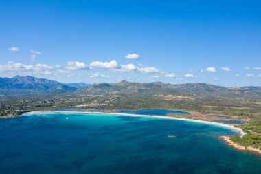 Spiaggia Lu Impostu 'nun havadan görünüşü. Süpürülen beyaz kumlu kıyı şeridi, canlı turkuaz suyu ve bulutlarla kaplı parlak mavi gökyüzünün altındaki engebeli dağlar..