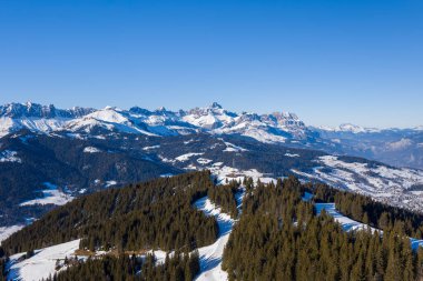 Fransız Alpleri 'ndeki Megeve yakınlarında açık mavi bir gökyüzünün altında dramatik dağ zirveleri bulunan, sık sık yeşil ormanlarla çevrili, kar kaplı kayak yamaçlarını süpüren dağlık arazi..