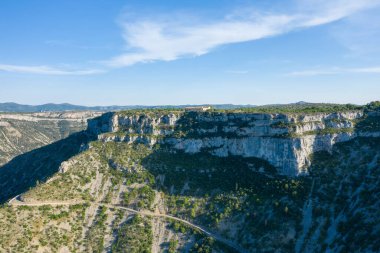 Dramatik kireçtaşı uçurumlarının havadan görünüşü ve Navacelles Sirki 'nde eğimli bir dağ yolu. Parlak güneş ışığı, berrak mavi gökyüzünün altındaki engebeli arazide kıtır kıtır gölgeler oluşturur..