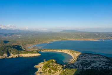 Yunanistan 'ın Navarino kenti yakınlarındaki yeşil tepeler ve açık kırsal bölgelerle çevrili turkuvaz suyu ve sakin bir lagünün bulunduğu hilal şeklinde kumlu bir körfezin havadan görünüşü açık mavi bir gökyüzü altında.
