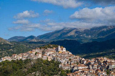 İtalya 'nın Rivello kentinin büyüleyici manzarası, kümelenmiş pastel evler ve dramatik dağlarla çevrili yeşil bir yamaçta tünemiş beyaz bir kilise ve dağınık bulutlu bir gökyüzü. Parlak gün ışığı dokuları ve doğal manzarayı güçlendirir..