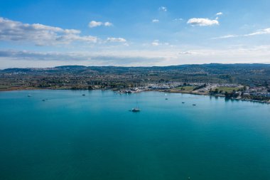 Yunanistan 'ın Kilada kenti yakınlarındaki huzurlu bir marina ve turkuaz körfezin havadan manzarası. Sakin su dağınık yelkenlileri yansıtır, dalgalı tepeler ve arka planda parçalı bulutlu bir gökyüzü.