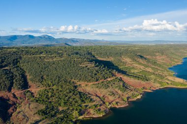 Engebeli kızıl toprak kayalıkları ve Salagou Gölü 'nün derin mavi sularını çevreleyen yoğun çam ormanı, uzak dağlar ve arka planda bulutlarla kaplı parlak bir gökyüzü..