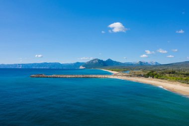 Canlı mavi Akdeniz suları Orosei kıyı şeridi boyunca uzun taştan bir dalgakıran ve kumlu bir plajla karşılaşıyor. Parlak, bulutlu bir gökyüzünün altında uzak dağlar var..