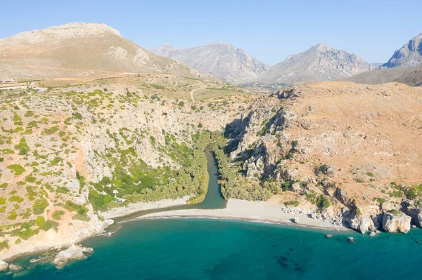 Bereketli yeşillikleri kesen kıvrımlı bir nehri olan Preveli sahilinin, dik kayalıklar ve parlak güneş ışığı altındaki kurak tepelerle çevrili havadan bakış açısı. Açık turkuaz deniz, kumlu bir kıyı şeridiyle buluşarak çarpıcı bir doğal karşıtlık yaratır..