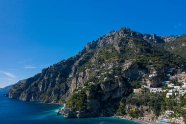 İtalya 'nın Positano kentindeki Amalfi Sahili' nin turkuaz sularından yükselen engebeli kireçtaşı dağları. Beyazlatılmış evler dramatik uçurumlara dağılmış durumda, canlı mavi gökyüzünün altında yemyeşil bir yer ile çevrili..