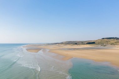 Plage de la Vieille eglise 'de sığ gel-git havuzları ve yumuşak dalgaları olan geniş kumlu bir sahil şeridine geniş bir hava perspektifi. Açık mavi gökyüzü ve uzak çimenli kum tepeleri sakin, açık bir kıyı manzarası yaratır..
