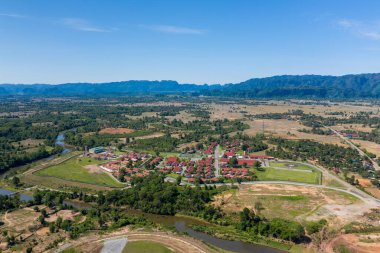 Kırsal bir vadide, kıvrımlı bir nehrin kenarına kurulmuş kırmızı çatılı evleri olan bir köyün hava manzarası. Sahnede açık alanlar, dağınık ağaçlar ve açık mavi gökyüzünün altında uzak dağ sırtları yer alıyor.