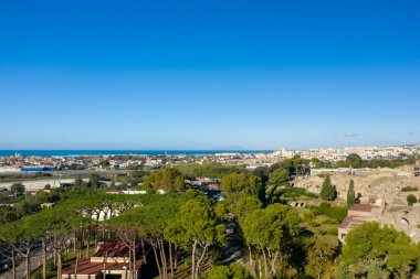 Eski harabeleri, yemyeşil yeşillikleri ve açık bir gökyüzü altında mavi Tyrhenian Denizi 'ne doğru uzanan modern şehirle Pompeii' nin geniş bir manzarası. Kompozisyon, güneydeki arkeolojik dokular, Akdeniz ağaçları ve kentsel gelişimi harmanlıyor.