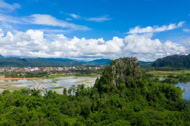Laos, Vang Vieng 'in eteklerindeki yemyeşil orman ve pirinç tarlalarının üzerinde çarpıcı bir kireçtaşı oluşumu yükseliyor. Sahne, dağınık bulutlarla dolu canlı bir mavi gökyüzünün altında geçiyor. Islak havada dramatik karst manzarasını yakalıyor.