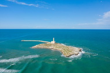 Panoramik bir hava perspektifi, İtalya 'nın Vieste kıyısındaki kayalık bir adada, turkuaz Adriyatik suları ve geniş bir mavi gökyüzü ile çevrili Santa Eufemia deniz fenerini yakalar. Sakin deniz manzarası engebeli kıyı şeridini vurguluyor.