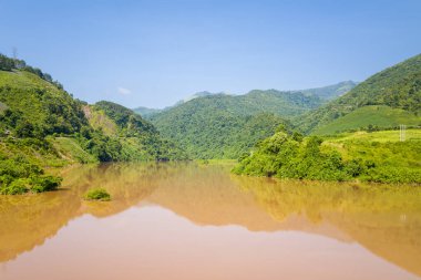 Çamurlu nehir, Kuzey Vietnam 'daki açık mavi gökyüzünün altındaki yemyeşil tepeleri ve yoğun bitki örtüsünü yansıtır. Parlak güneş ışığı uzak manzaranın doğal dokularını ve sakin atmosferini vurgular.