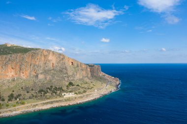 Monemvasias 'ın engebeli turuncu kayalıkları ve açık gökyüzünün altında açık mavi Akdeniz' i çevreleyen kıvrımlı bir yol. Manzara, dokulu kaya yüzeyleri, seyrek bitki örtüsü ve sakin sulara sahiptir..