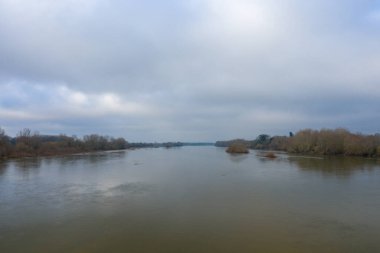 Loire nehrinin yapraksız ağaçlar ve çalılarla çevrili geniş manzarası, soluk kış bulutlarının altında solgun yansımalar. Sakin su ve bastırılmış renkler dingin ve düşünceli bir ruh halini çağrıştırıyor..