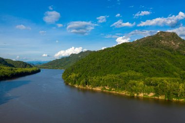 Parlak güneş ışığı yoğun yeşil tepeleri aydınlatır geniş Mekong nehri üzerinde yükselen, dağınık bulutlar parlak mavi gökyüzünü noktalar. Manzara, verimli tropikal bitki örtüsü, hafif eğimli arazi ve sarmaş dolaş sakin bir su yoluna sahiptir.