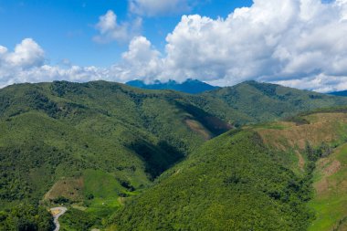Yuvarlanan yeşil tepelerin geniş manzarası ve canlı mavi gökyüzünün altındaki orman yamaçları Phou Khoun ve Luang Prabang, Laos kırsallarındaki dramatik kümülüs bulutlarıyla dolu. Güneş ışığı ve gölgeler dalgalanan dalgalar boyunca dinamik desenler oluşturur.