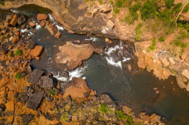 Havadan bakıldığında, Laos 'un güneyindeki Tad Tayicsua yakınlarında dağılmış kaya parçaları, açıkta kalmış taş ve yeşillik parçaları olan engebeli bir nehir yatağı görülüyor. Güneş ışığı dokuları, topraksı portakal kayaları ve karanlık su arasındaki zıtlığı vurgular ve çarpıcı bir şekilde