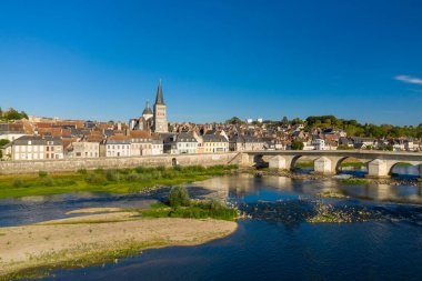 La Charite sur Loire 'ın panoramik hava perspektifi, tarihi taş köprüsü, ortaçağ manastır kulesi ve Loire nehri boyunca geleneksel evleri yer almaktadır. Parlak mavi gökyüzü ve yemyeşil nehir kıyısı bitki örtüsü bu sakin karakteri vurgular.