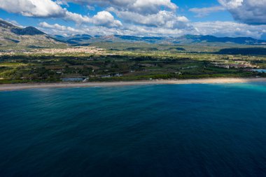 Derin mavi suların havadan görünüşü kumlu bir kıyı şeridiyle buluşuyor, yeşil kırsal ve uzak dağ sıraları Orosei, Sardunya yakınlarındaki dramatik bulutların altında..