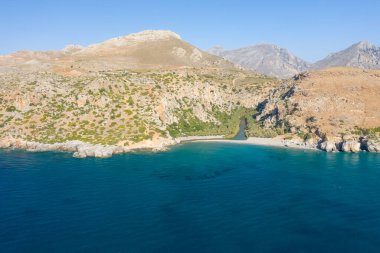 Girit 'in Preveli kenti yakınlarındaki gözlerden uzak bir körfez manzarası canlı turkuaz su, kumlu kıyı şeridi ve seyrek Akdeniz bitkileriyle kaplı dik kayalık uçurumlar. Parlak güneş ışığı ve berrak mavi gökyüzü sakin bir kıyı manzarası yaratır..