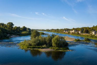 Sakin bir nehir sahnesinde, sakin sularla çevrili, La Charite sur Loire 'deki nehir kıyısı boyunca uzanan evler ve yemyeşil ağaçlarla çevrili küçük yeşil bir ada bulunur. Açık mavi gökyüzü ve yumuşak ışık huzur dolu bir Loire Vadisi manzarasını canlandırıyor..