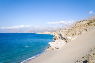 Expansive sandy slope descends to rugged limestone cliffs overlooking deep blue Mediterranean waters near Agios Pavlos, Crete. The scene is sunlit and open, with distant mountains under a clear sky.