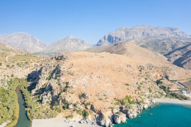 Girit, Preveli 'de, canlı turkuaz bir sahil şeridiyle buluşan güneşli kayalık tepelerin geniş hava manzarası. Manzarada engebeli uçurumlar, yeşilliklerle çevrili dolambaçlı bir nehir ve berrak mavi gökyüzünün altındaki uzak dağ zirveleri yer alıyor..
