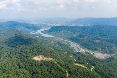 Hava görüntüsü, yoğun yeşil orman, dolambaçlı yollar ve geniş nehir vadisi içinde bir hidroelektrik tesisatı ve dairesel yapıyı gösteriyor. Sahne, modern altyapı ile çevreleyen tropikal dağ bölgesi arasındaki zıtlığı yakalıyor.